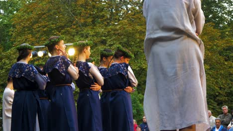 Adult-caucasian-female-dancers-in-traditional-blue-folk-costumes-perform-in-a-dance-performance-in-open-air,-sunny-summer-evening,-happy,-Latvian-national-culture,-medium-handheld-shot