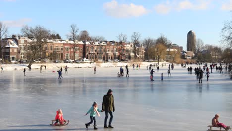 Urban-picturesque-scenery-of-people-on-frozen-canal-in-city-environment-with-winter-barren-trees-and-white-snow-in-cold-cozy-landscape-against-a-blue-sky