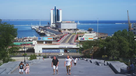 Establishing-Shot-of-Tourists-Walking-Up-Potemkin-Stairs-with-Odessa-Seaport-Terminal-in-Background-on-Summers-Day,-Odessa,-Ukraine