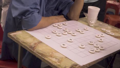 Elderly-Men-In-Mask-Playing-Chinese-Chess-In-Chinatown,-Singapore-During-Their-Free-Time