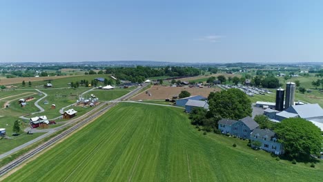 Eine-Luftaufnahme-Einer-Dampfmaschine,-Die-Rauch-Und-Dampf-Bläst,-Mit-Personenwagen,-Die-Sich-An-Einem-Schönen-Wolkenlosen-Frühlingstag-An-Einem-Maislabyrinthpark-Und-Einer-Ackerlandlandschaft-Vorbei-Nähern