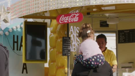 Parent-holding-child-on-shoulders-as-they-get-snack-from-a-vendor-at-a-outdoor-Balvarian-festival-at-Stuttgart,-Germany,-Europe-at-sunset,-panning-view-angle