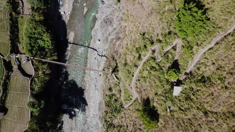 Gebirgiger-Zick-Zack-Schotterweg,-Der-Zur-Hängebrücke-Aus-Metall-Führt,-Flussüberquerung,-Smaragdgrünes-Bergwasser,-Graue-Felsen,-Grüne-Bäume,-Von-Oben-Nach-Unten-Absteigend,-Vogelperspektive