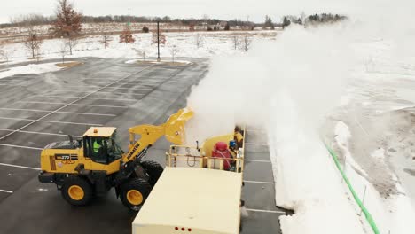 Luftaufnahme-Eines-Schneepflugtraktors,-Der-Einen-Eimer-Voller-Schnee-In-Die-Schneeschmelzmaschine-Wirft