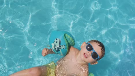 Cool-boy-looking-to-camera,-wearing-sunglasses-and-floaties-Pristine-water-pool,-Topdown