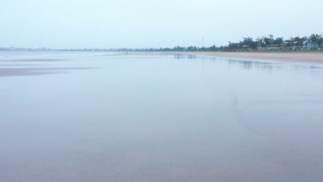 Plano-De-Tierra-Que-Viaja-De-La-Costa-Tranquila-Y-Acuosa,-El-Dron-Se-Eleva-Para-Mostrar-La-Playa,-El-Cielo-Suave-Y-Sombrío,-Una-Sola-Persona-En-La-Playa,-árboles-En-La-Distancia,-Ambiente-Tranquilo-Y-Tranquilo,-Playa-Vacía,-Cielo-Nublado,-Pacífico