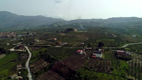 Aerial-overview-over-beautiful-farmlands-with-olives-and-oranges-crops-been-cultivated-in-Sicily,-Italy-over-hilly-terrain-at-daytime-with-the-view-of-small-village-in-distance