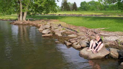 Eine-Atemberaubende-Seelandschaft-Mit-Kristallklarem-Grünem-Wasser-Und-Ein-Mädchen,-Das-Am-Ufer-Des-Sees-Sitzt-Und-Mit-Kieselsteinen-Spielt