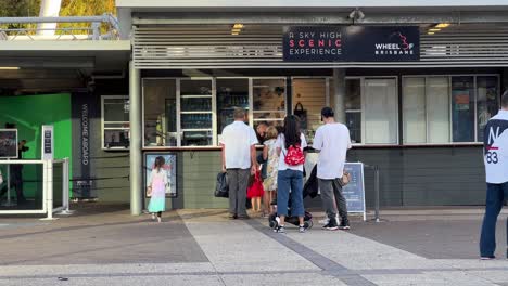 Happy-tourists-lining-up-at-the-ticket-counter-to-go-on-gondola-ride-on-channel-seven-wheel-of-Brisbane,-iconic-landmark-ferris-wheel-scenic-experience-at-South-bank-parklands,-Queensland,-Australia