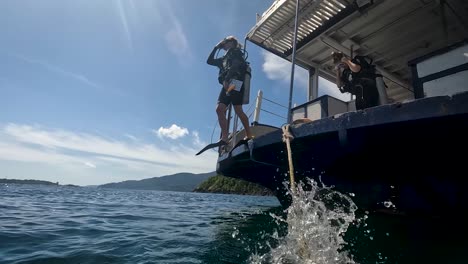 Tiro-En-ángulo-Bajo-Con-Un-Hombre-Vestido-Con-Traje-De-Neopreno-Y-Aletas-Mientras-Se-Sumerge-En-El-Mar-Desde-El-Borde-Del-Barco-Seguido-Por-Otros-Turistas-En-Un-Día-Soleado