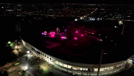 Vista-Aérea-De-Un-Concierto-En-Vivo-Iluminado-En-Un-Estadio-En-Medio-De-Una-Ciudad-Por-La-Noche