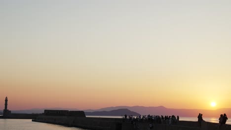 Hermosa-Vista-De-La-Puesta-De-Sol-Sobre-El-Mar-Egeo-En-Chania,-Turistas-Disfrutando-Del-Antiguo-Puerto-Veneciano-Con-Un-Faro-Histórico-Durante-La-Noche-De-Verano,-Lugar-Turístico-En-Grecia