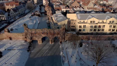 Paisaje-De-Nieve-De-Invierno-Aéreo-Congelado-Sobre-El-Río-Berkel-Con-Portal-Medieval-Berkelpoort-De-La-Ciudad-Histórica-Holandesa-Zutphen-En-Los-Países-Bajos-En-El-Fondo