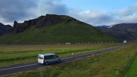 Van-Fährt-Auf-Asphaltstraße,-Vorbei-An-Grünen-Feldern-Mit-Blick-Auf-Die-Berge-In-Island