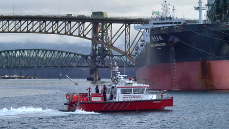 Barco-De-Bomberos-Navegando-Por-El-Río-Con-Un-Gigantesco-Barco-Portacontenedores-Y-Un-Puente-Con-Coches-De-Conducción-En-Segundo-Plano