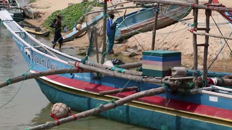 Cerca-De-Un-Hermoso-Barco-De-Color-Azul-Con-Franja-Roja-Anclado-En-La-Costa-Del-Mar,-Mientras-La-Gente-Se-Mueve-Haciendo-Su-Trabajo-Cerca-De-él