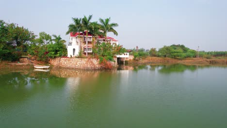 Aerial-shot-traveling-backward-of-a-white-tropical-house-with-balcony-and-red-roof-surrounded-by-palm-trees,-a-boat-and-green-plants