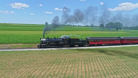 Eine-Luftaufnahme-Einer-Dampfmaschine,-Die-Rauch-Und-Dampf-Ausstößt,-Während-Reisezugwagen-An-Einem-Wunderschönen-Wolkenlosen-Frühlingstag-Auf-Einer-Eingleisigen-Strecke-Durch-Bepflanzte-Felder-Und-Ackerland-Fahren