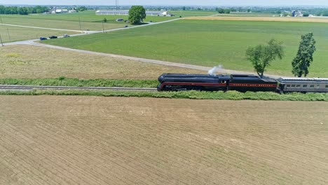 Una-Vista-Aérea-De-Un-Motor-De-Vapor-Que-Sopla-Humo-Y-Vapor-Con-Autocares-De-Pasajeros-Que-Viajan-En-Una-Sola-Vía-A-Través-De-árboles-Y-Campos-De-Cultivo-En-Un-Hermoso-Día-De-Primavera-Sin-Nubes