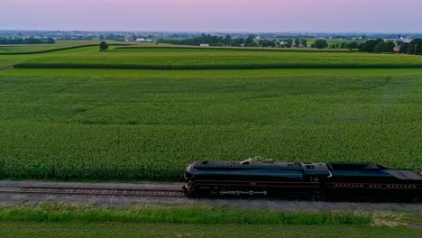 Una-Vista-Aérea-De-Un-Tren-De-Vapor-Que-Se-Acerca-Volando-Viajando-A-Través-De-Tierras-De-Cultivo-Y-Campos-De-Maíz-Que-Soplan-Humo-En-Un-Día-Soleado-De-Verano