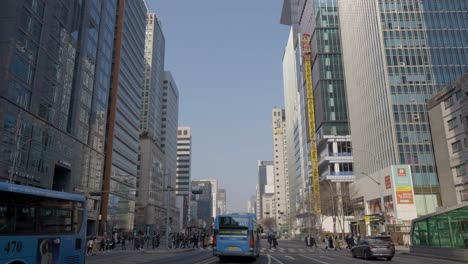 Busy-urban-city-street-life-with-pedestrians-crossing-road-by-buses-and-traffic-daytime