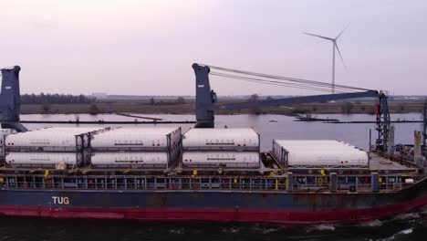 Aerial-Starboard-Side-View-Of-Pacific-Fortune-Cargo-Ship-Passing-By-With-Still-Wind-Turbine-In-background