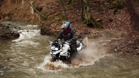 Motocicleta-Todoterreno-Cruzando-El-Río-Salpicando-Agua-Y-Lodo-Sucio-A-Cámara-Lenta,-Prueba-De-Deporte-Extremo-Potente-Ciclista-Fumando-Motor-En-El-Húmedo-Bosque-Otoñal