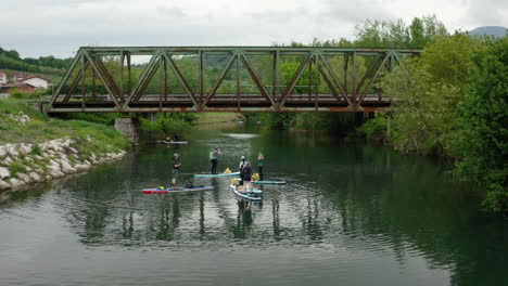 Toma-Más-Amplia-De-Drones-De-Personas-Remando-Y-Descansando-En-Un-Río-Verde-En-Eslovenia-Con-Un-Viejo-Puente-De-Tren-De-Hierro-Y-Oxidado-En-El-Fondo