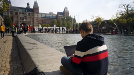 Hübscher-Junger-Mann-Sitzt-Vor-Dem-Rijksmuseum-Mit-Einem-Amsterdam-Schild-Und-Tippt-In-Ein-Notizbuch