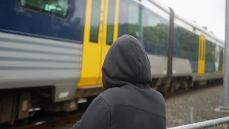 Cámara-Lenta-De-Un-Hombre-Rubio-Con-Una-Capucha-Negra-De-Pie-Y-Mirando-Un-Tren-Que-Pasa-Por-Los-Ferrocarriles,-En-Un-Día-Nublado