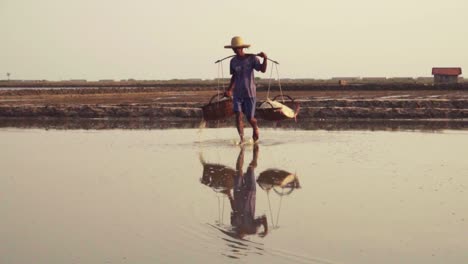 Zeitlupe-Eines-Arbeiters,-Der-Mit-Wasser-Tropfende-Salzkörbe-Auf-Den-Reflektierenden-Salzfeldern-Asiens-Trägt