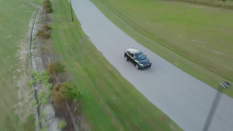 Vista-Aérea-De-Un-Coche-Cruzado-Verde-Que-Se-Aleja-Por-Una-Carretera-Rural-Vacía