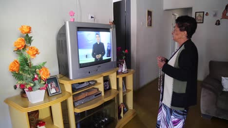 Una-Mujer-Sostiene-Su-Rosario-Mientras-Reza-Frente-A-Un-Televisor-Escuchando-Al-Sacerdote