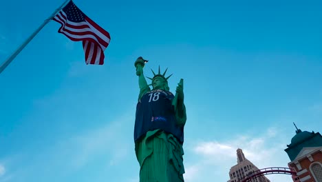 Toma-Panorámica-De-La-Estatua-De-La-Libertad-Con-Una-Camiseta-De-Baloncesto-Y-La-Bandera-Estadounidense-Ondeando-En-El-Viento-Fuera-Del-Hotel-Y-Casino-De-Nueva-York-En-Las-Vegas,-Nevada,-Estados-Unidos