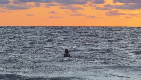 Surfer-on-surfboard-paddling-over-waves-near-the-Baltic-sea-Karosta-beach-at-Liepaja-during-a-beautiful-vibrant-sunset-at-golden-hour,-medium-shot-from-a-distance