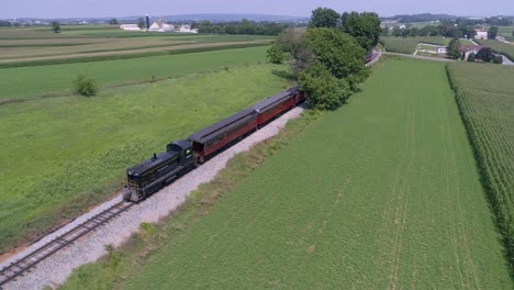 Una-Vista-Aérea-De-Una-Locomotora-Diésel-Tirando-De-Coches-De-Pasajeros-Antiguos-A-Través-De-La-Campiña-Amish