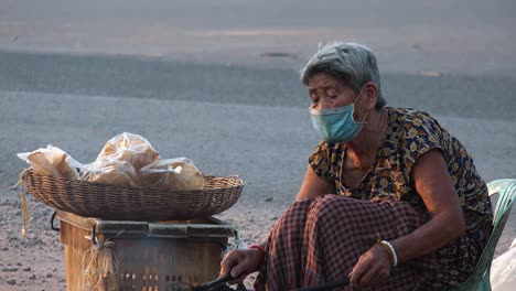 Plano-Medio-De-Una-Dama-Con-Una-Máscara-Facial-Usando-Palos-Largos-Cocinando-Pan-Plano-Sobre-Un-Fuego-Humeante-En-El-Lado-De-La-Carretera-Mientras-Las-Motocicletas-Pasan-En-El-Fondo