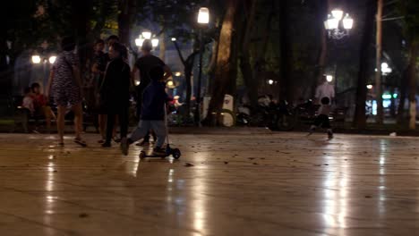 Kid-rides-glider-scooter-while-his-family-watches-from-a-nearby-bench-in-a-local-park-at-night,-Low-angle-tracking-handheld-shot