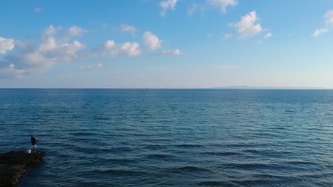 Plaka-beach-in-the-east-point-of-the-island-with-the-Vrahos-beach-and-snack-bar-sign-and-a-man-fishing-on-the-rocks,-Aerial-drone-flyover-shot