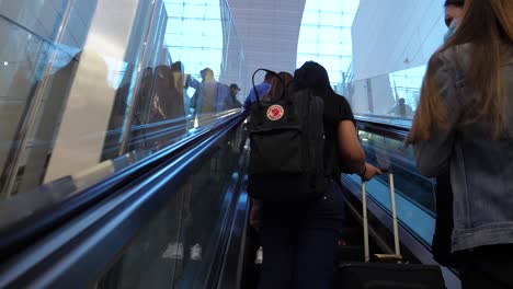 Mujer-Con-Mascarilla-En-El-Aeropuerto-En-Una-Escalera-Mecánica,-Viajando-Con-Personas-Durante-Una-Pandemia,-Virus-De-La-Corona