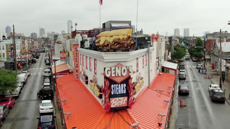 Geno&#39;s-Steaks,-Berühmtes-Philly-Cheesesteak-Mit-Der-Skyline-Von-Philadelphia-In-Der-Ferne-Im-Sommer,-Amerikanische-Flagge-An-Einem-Berühmten-Touristenstopp