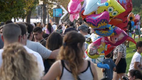 Young-boy-selling-floating-colourful-funny-balloons-in-crowded-park-full-of-tourists---visitors