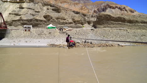 A-man-crossing-over-a-river-on-a-rope-bridge,-pulling-him-in-a-cart-to-the-other-side