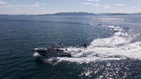 Wakesurfer-Detrás-De-Una-Lancha-Motora-Que-Acelera-Sobre-El-Agua-Azul-Oscuro-En-El-Lago-Flathead,-Kalispell,-Montana-En-Un-Día-De-Verano---Antena