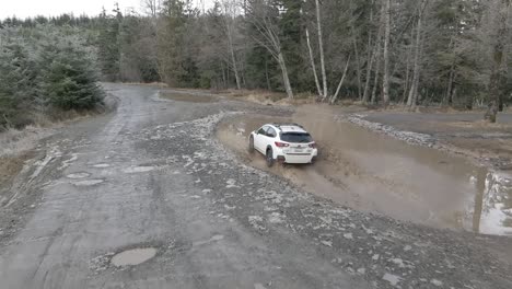 Ein-Einzelner-Weißer-Subaru-Crosstrek-Fährt-In-Eine-Tiefe-Schlammpfütze,-Aus-Der-Schmutziges-Wasser-Spritzt,-Luftaufnahme-In-Zeitlupe,-Illustrativer-Leitartikel