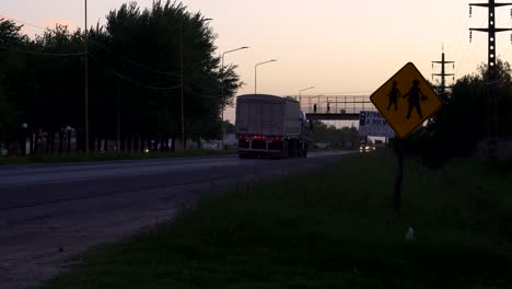 Tráfico-A-Lo-Largo-De-Una-Ruta-Al-Atardecer,-Con-Personas-De-Pie-En-Un-Puente-Peatonal-Distante