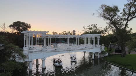 Aerial-pan-left-of-boats-sailing-under-white-bridge-in-Rosedal-gardens,-Palermo-neighborhood-at-sunset,-Buenos-Aires