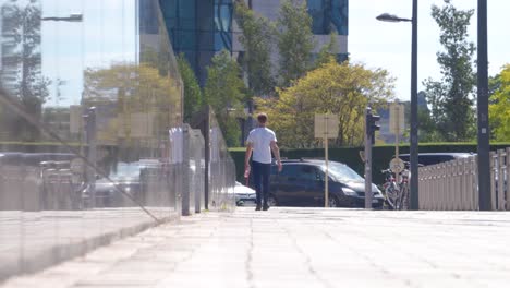 Toma-De-Mano-De-Un-Hombre-Caminando-En-La-Acera-Lejos-De-La-Cámara-Junto-A-La-Pared-Reflectante-En-Un-Día-Cálido-Y-Soleado-En-Bruselas,-Bélgica