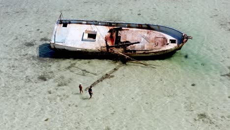 Luftaufnahme-Von-Zwei-Personen,-Die-Das-Rostende-Schiffswrack-Betrachten,-Das-Im-Seichten-Wasser-Des-Strandes-Liegt