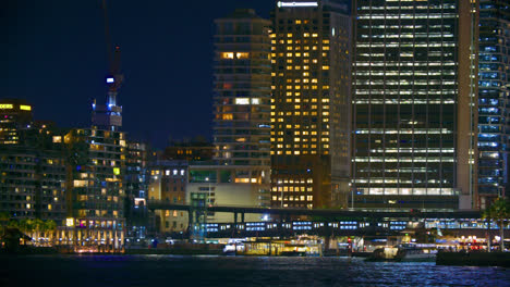 Tren-Que-Pasa-Por-Edificios-Frente-Al-Mar-Iluminados-Por-La-Noche-En-Sydney,-Nueva-Gales-Del-Sur,-Australia